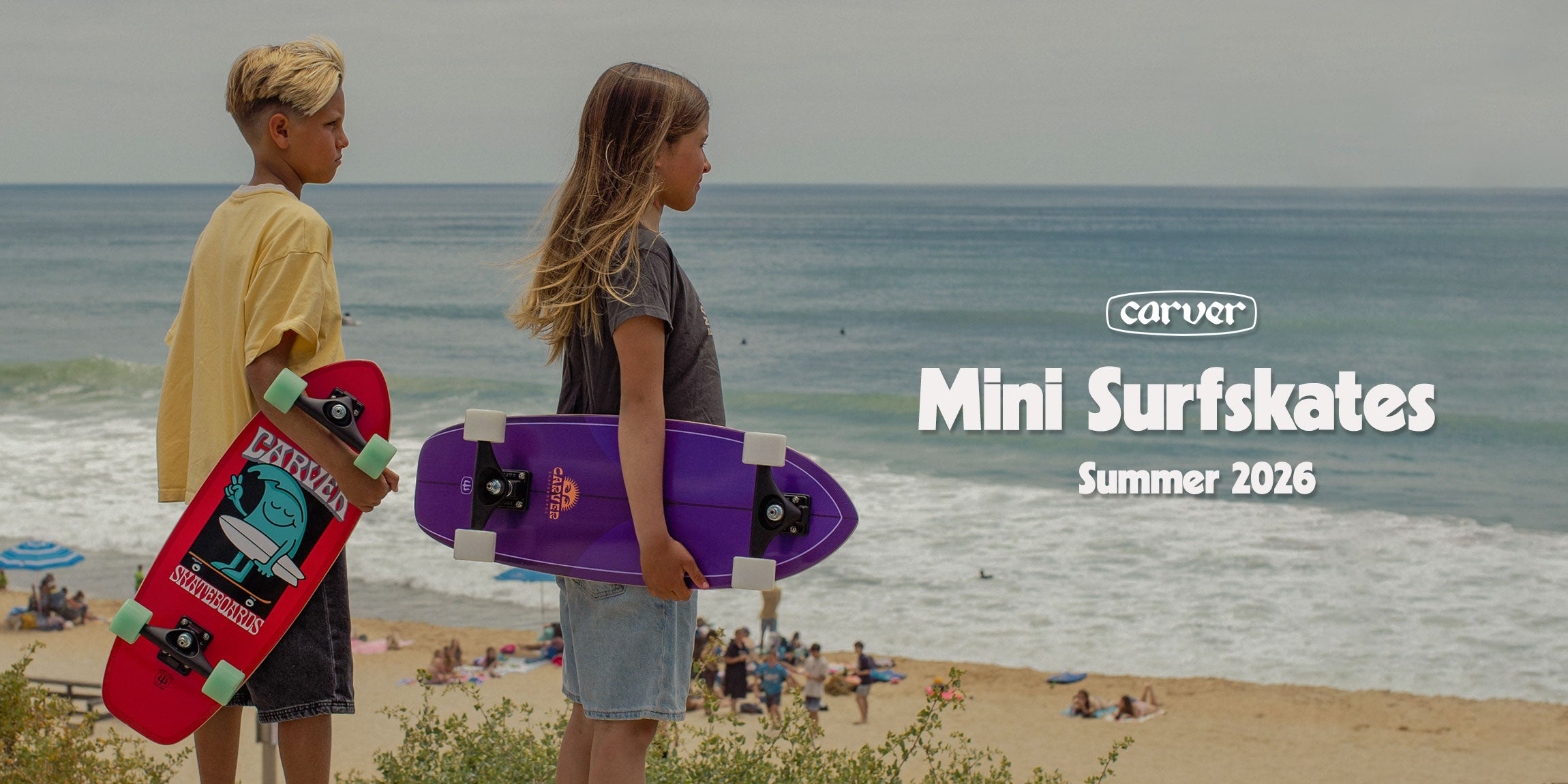 Two children holding Carver Wavy Davy and Purple Haze mini surfskates on a beach.