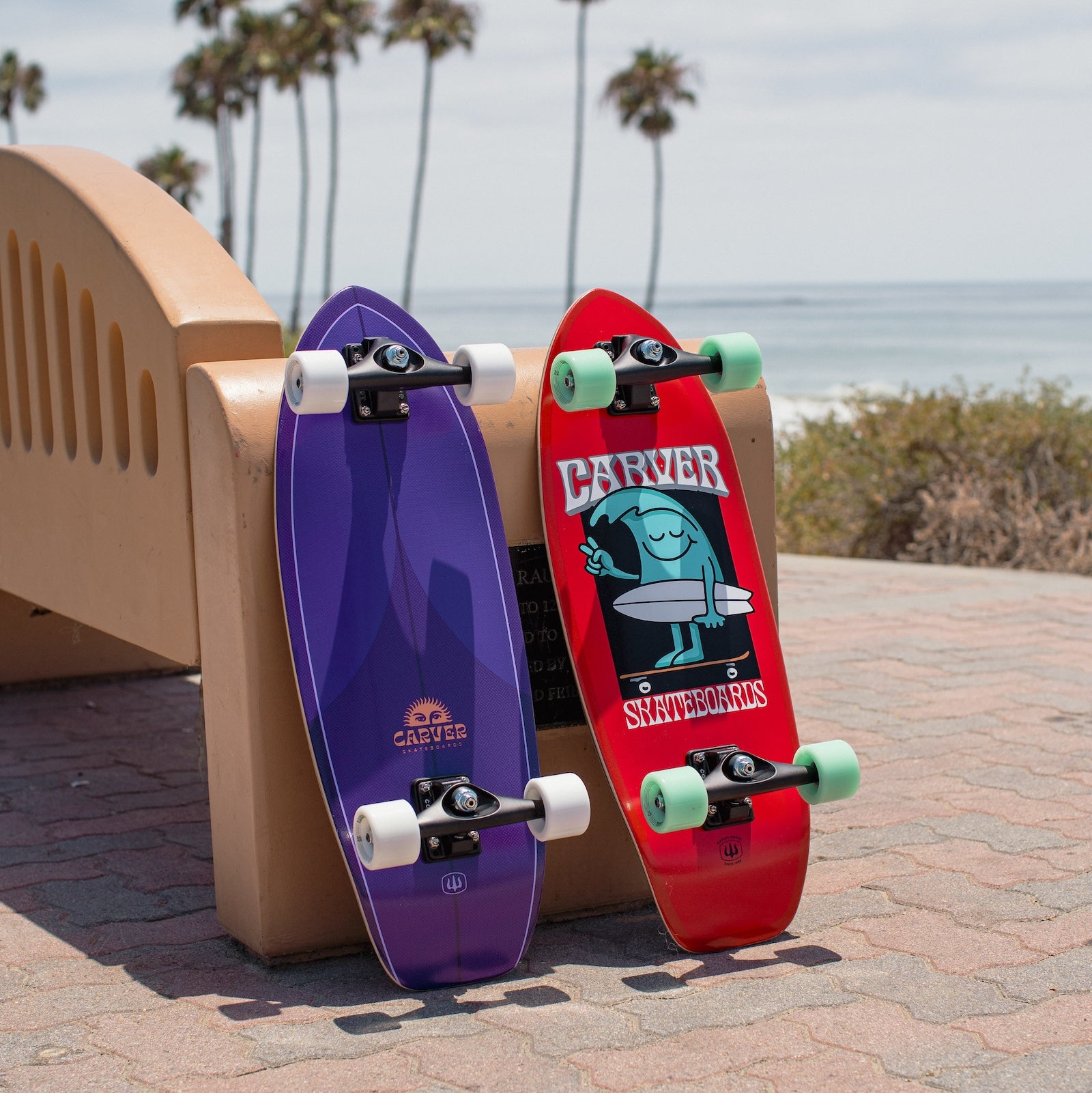 Two Carver surfskate skateboards, one purple (Purple Haze) and one red with a character design (Wavy Davy), leaning against a bench overlooking the beach.