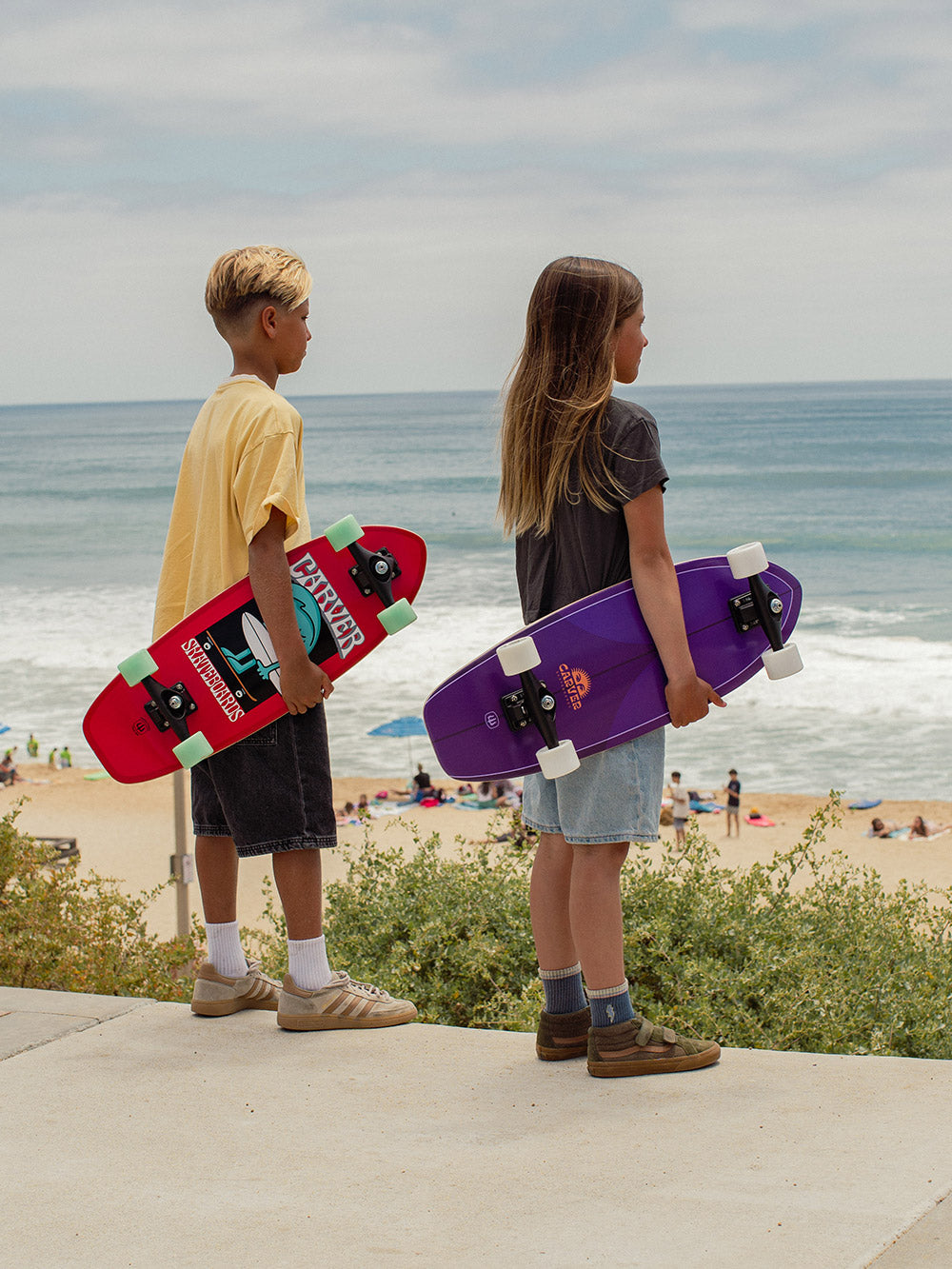 Two children holding Carver Wavy Davy and Purple Haze mini surfskates on a beach.