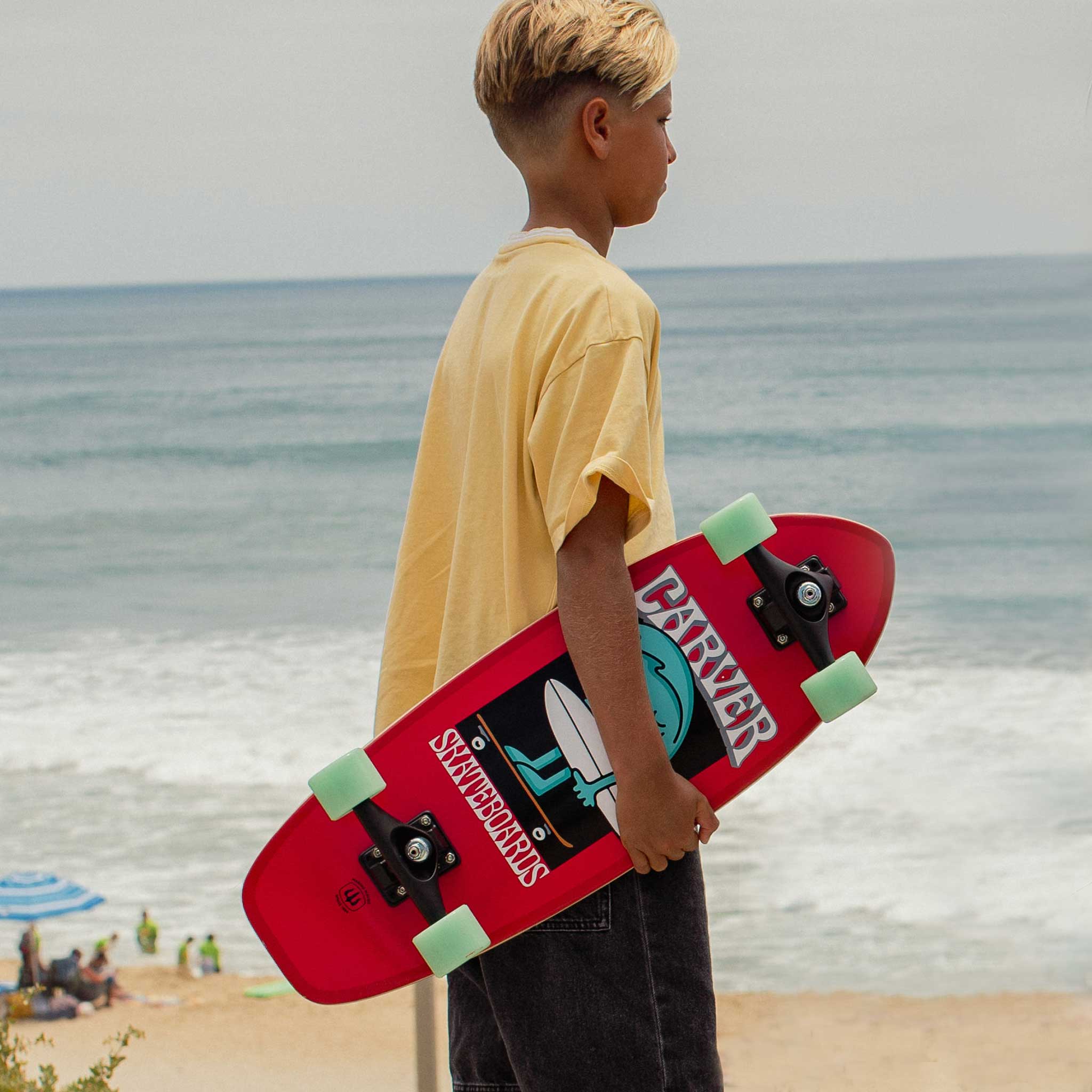 Child holding a Carver Wavy Davy mini surfskate with ocean and beach in the background