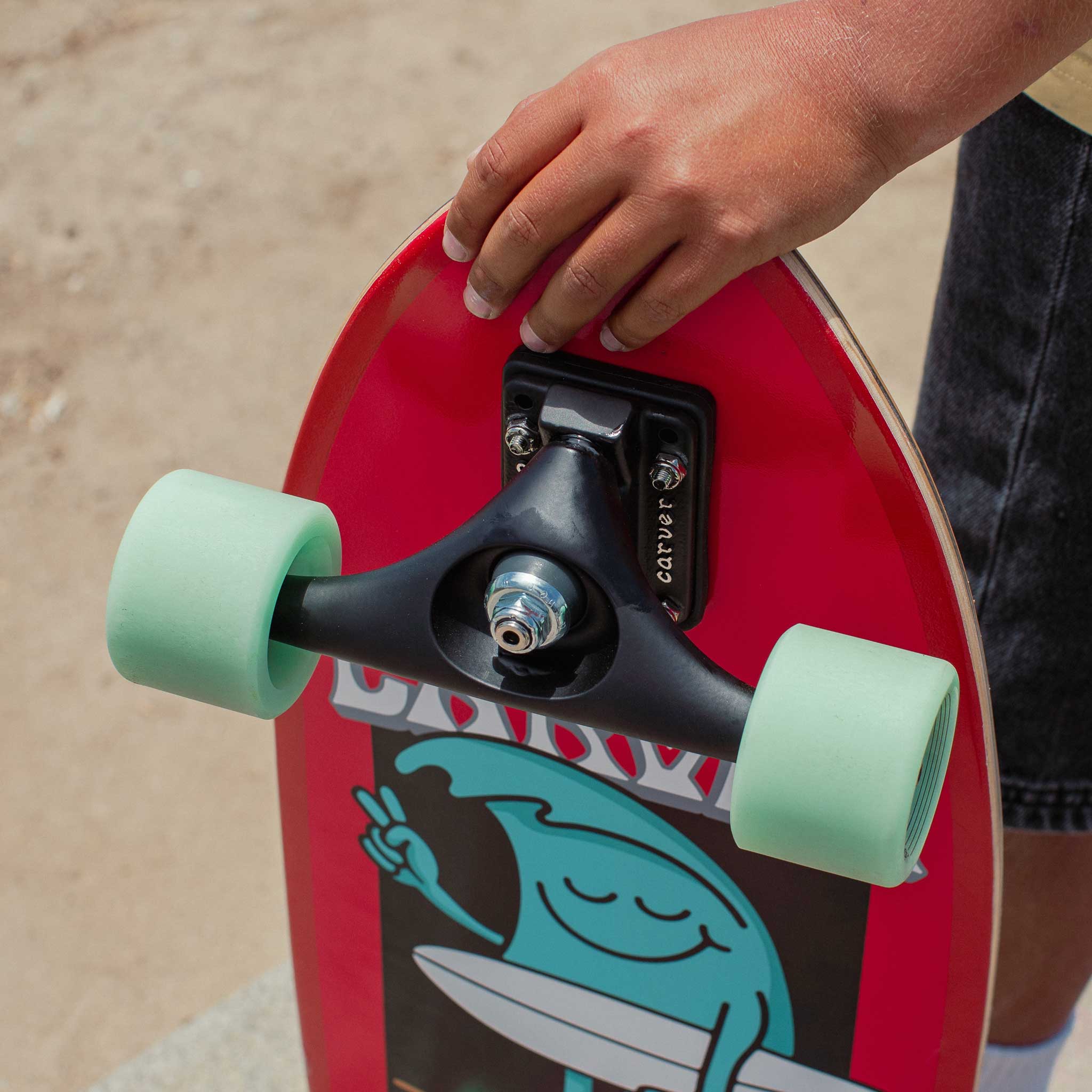 Red Carver Wavy Davy mini surfskate skateboard with green wheels held by a person on a blurred background