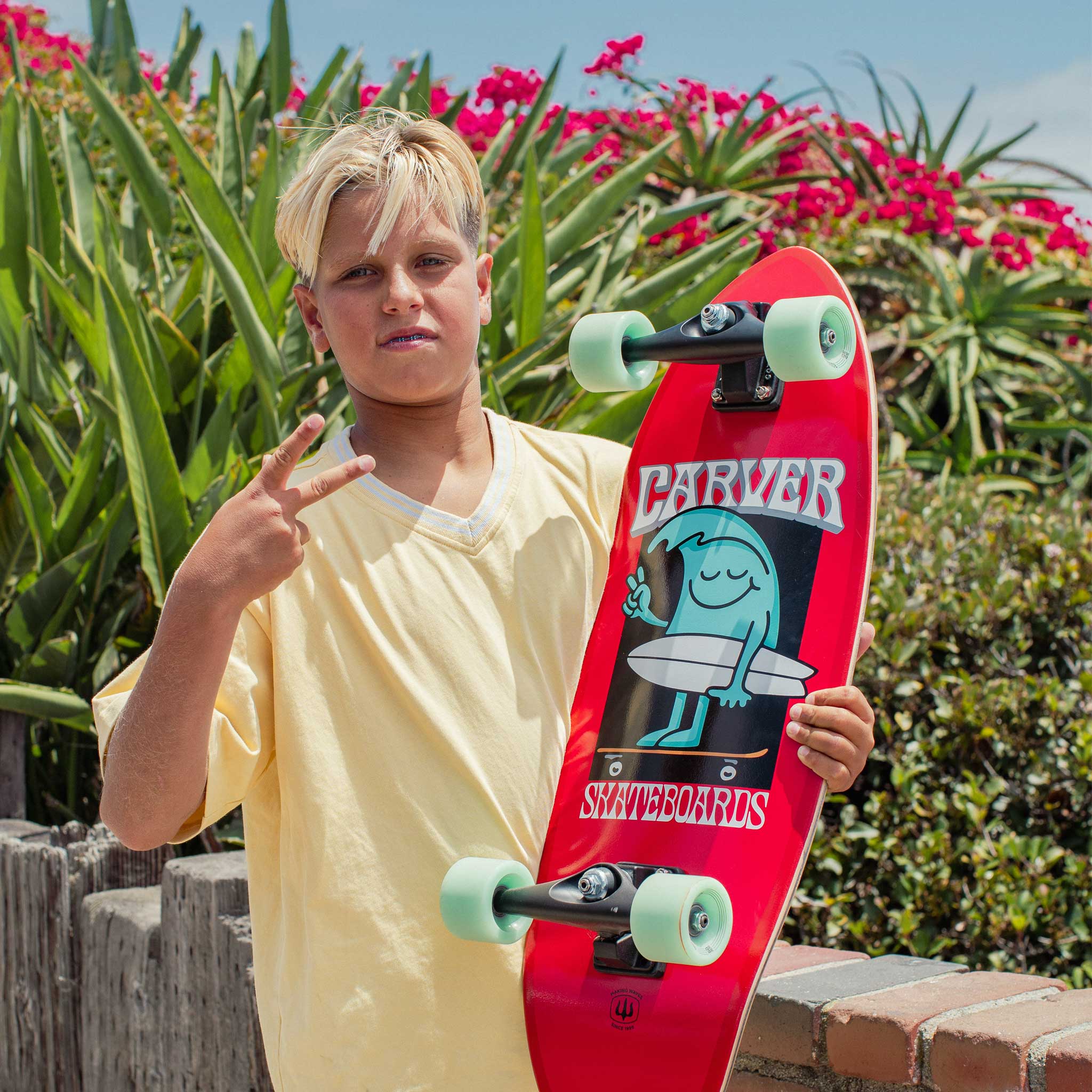 Person holding a Carver Wavy Davy mini surfskate, standing outdoors with greenery and flowers in the background.