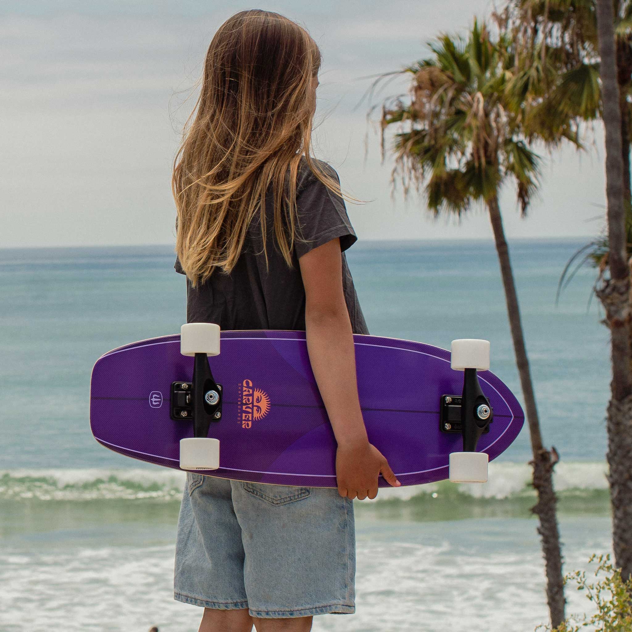 Person holding a purple Carver Purple Haze mini surfskate skateboard by the ocean with palm trees in the background