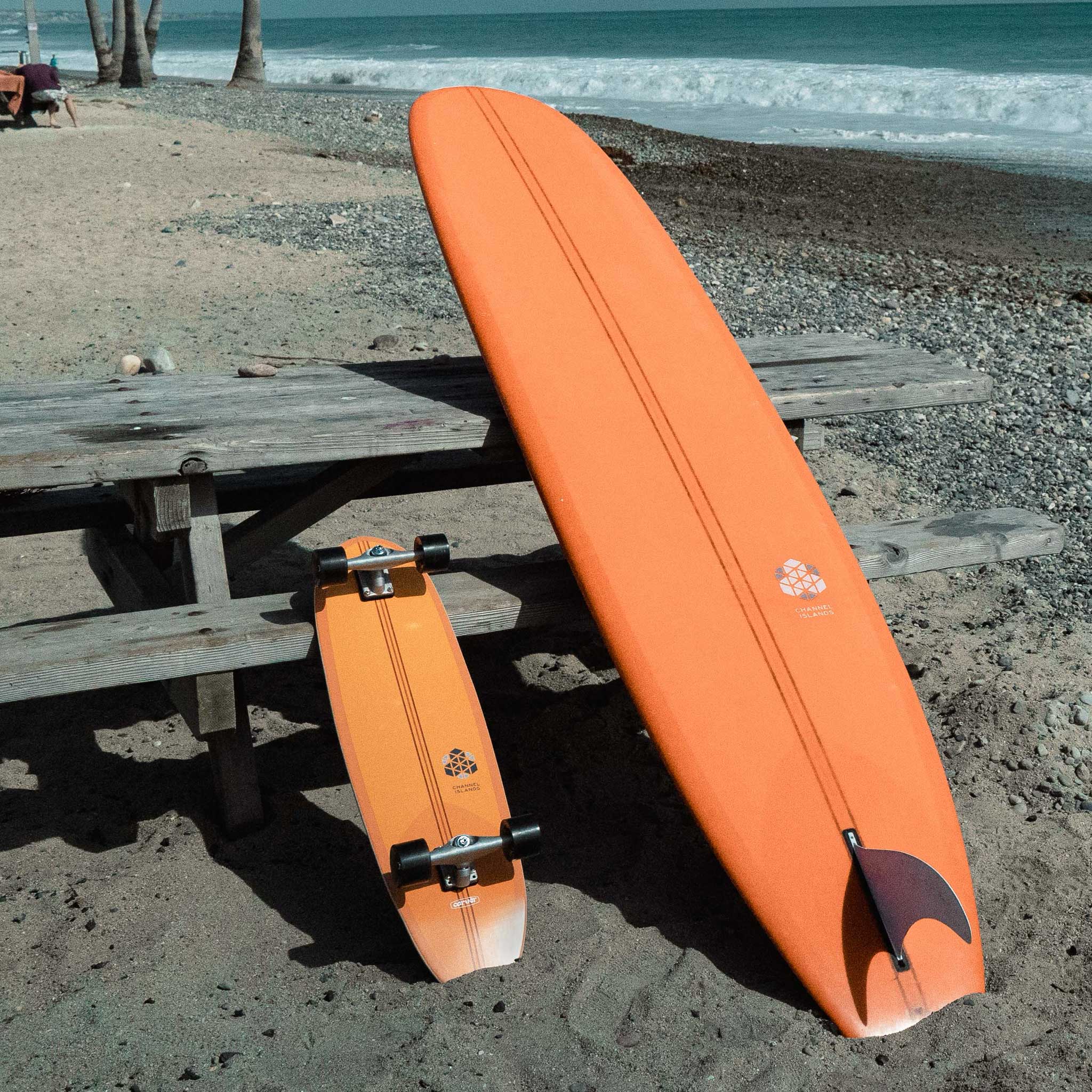 Two orange boards (Carver CI Noserider surfskate skateboard and Channel Islands Noserider surfboard) on a beach with a picnic table in the background.