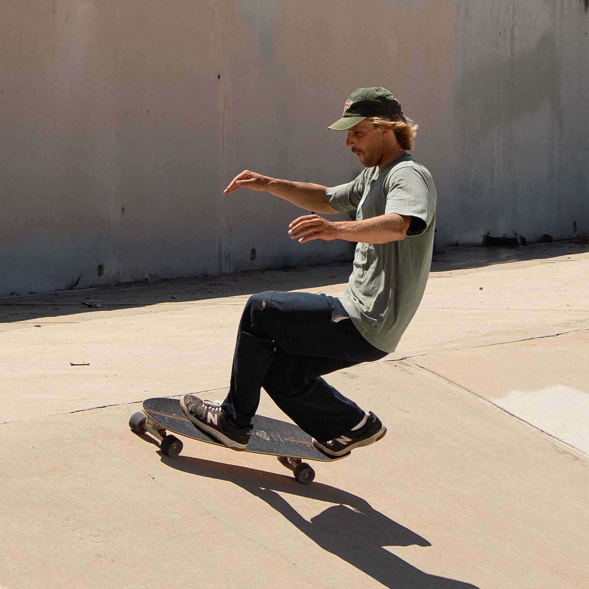 Person skateboarding on Carver CI Noserider surfskate on a concrete surface with a plain wall in the background