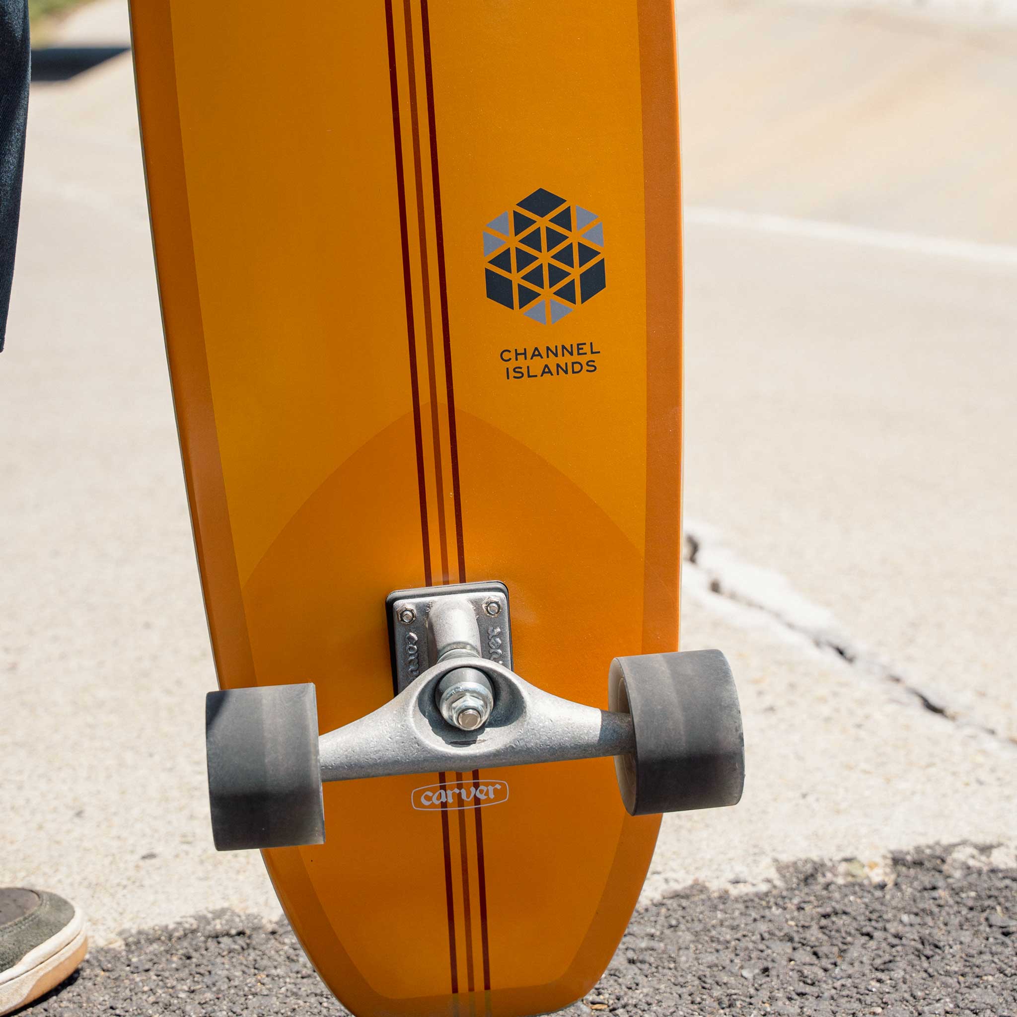 Close-up of an orange Carver CI Noserider surfskate skateboard with 'Channel Islands' branding on a concrete surface.