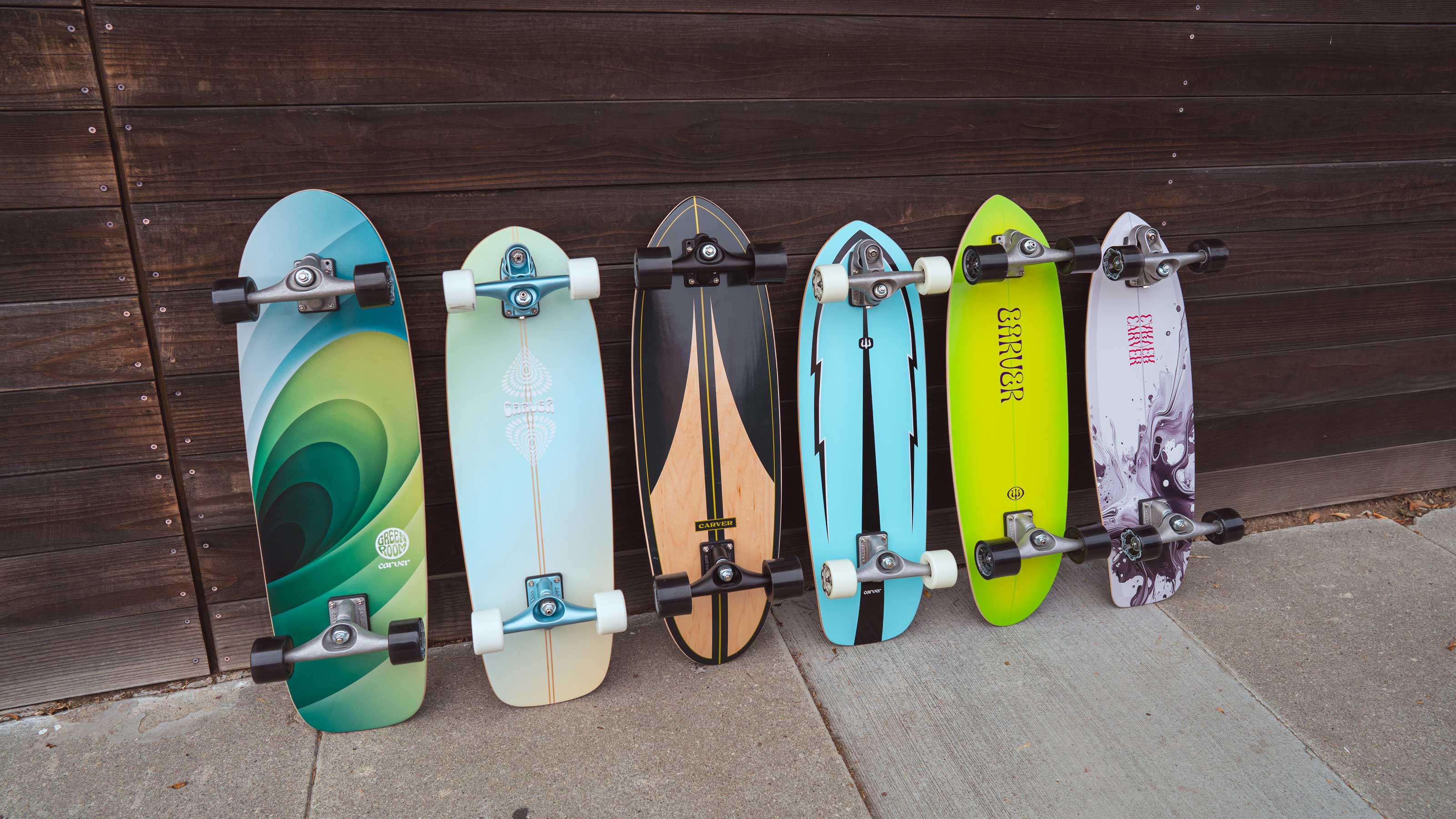 Six Carver surfskate skateboards with different colorful decks are lined up vertically against a dark brown wooden wall on a concrete sidewalk.