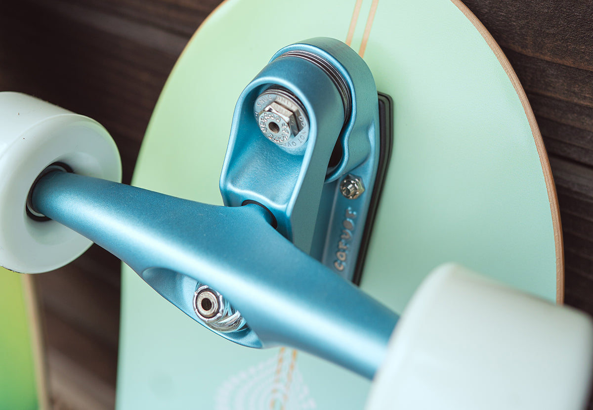 Close-up of a Carver C7 surfskate truck, showing a mint green Cosmic Haze deck with Impala Blue Metallic C7 trucks and white Roundhouse Slick wheels against a dark wood background.