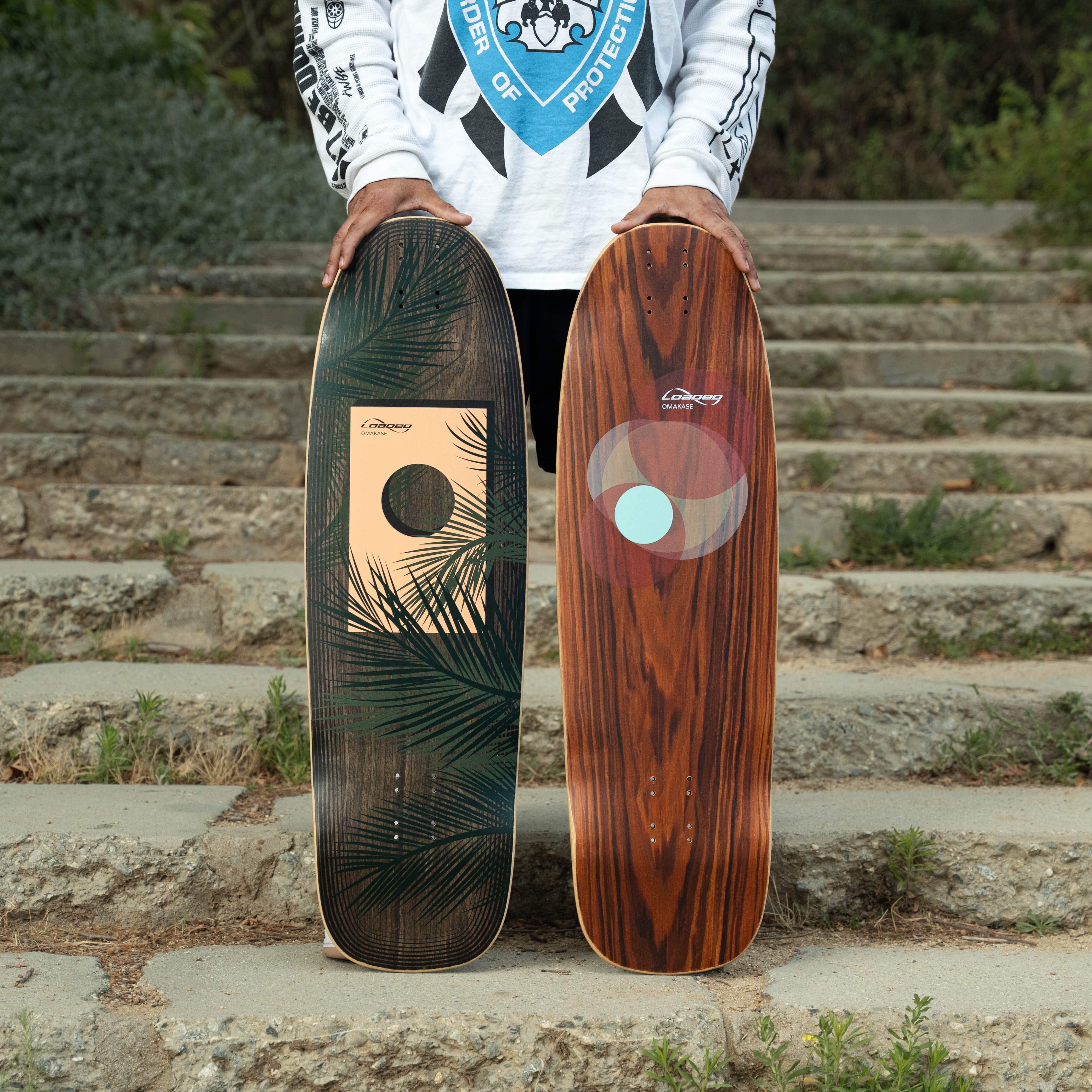 Two Loaded Omakase skateboard decks with Palm and Roe graphics held by a person on stone steps with a blurred background